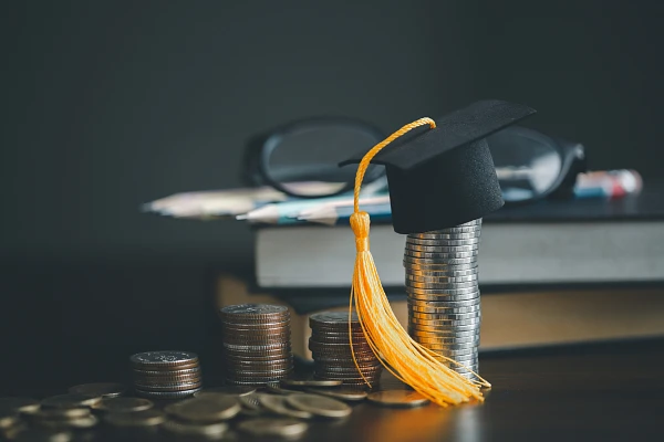 A graduation cap sits atop a stack of coins, surrounded by books, glasses, and more coins, symbolizing the cost of education.