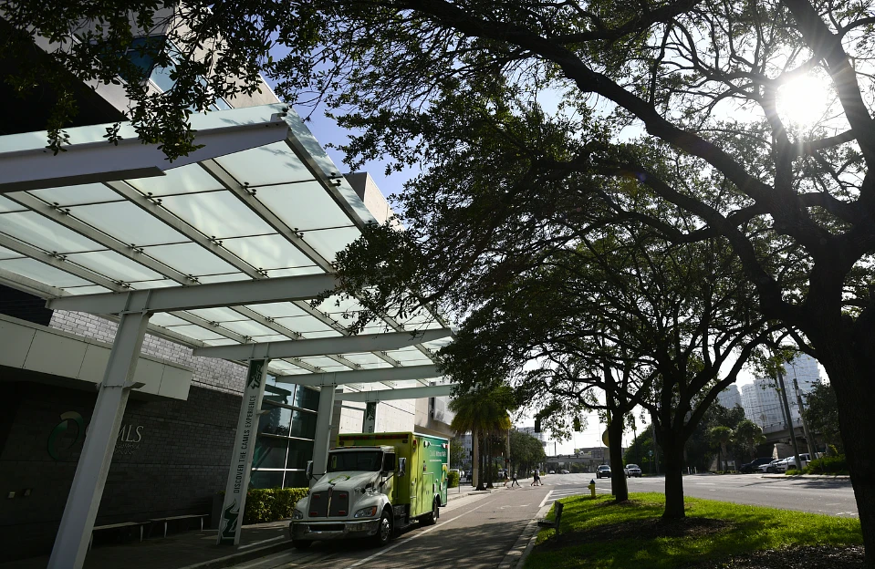 A green and white truck is parked under a modern glass canopy next to a tree-lined street on a sunny day.