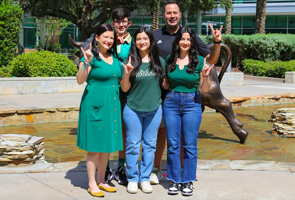 A group of five people in green and black attire stands smiling in front of a bull statue, making hand gestures outdoors.