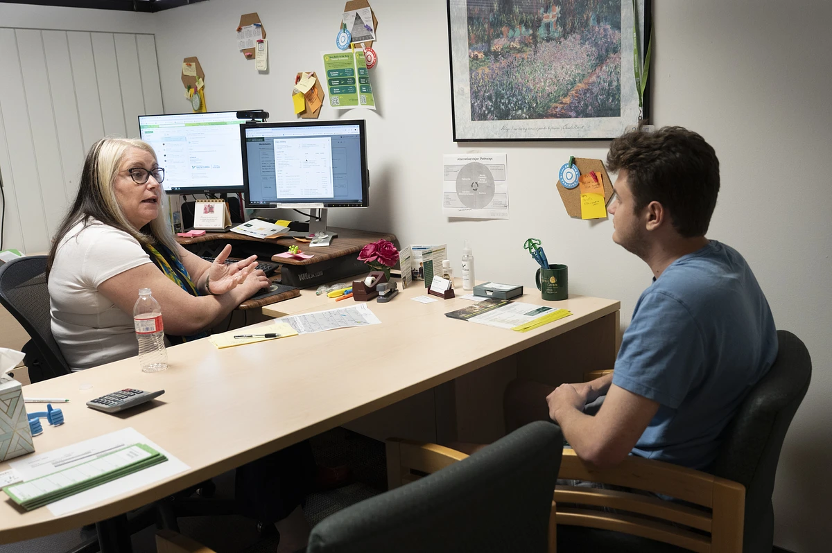 A woman is having a conversation with a man in an office setting while sitting across a desk from each other.
