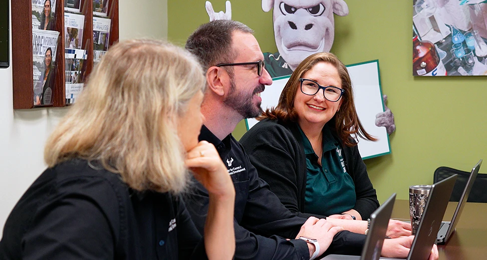 Three people are sitting around a table with laptops, talking and smiling, while a person in a cow costume gestures behind them.
