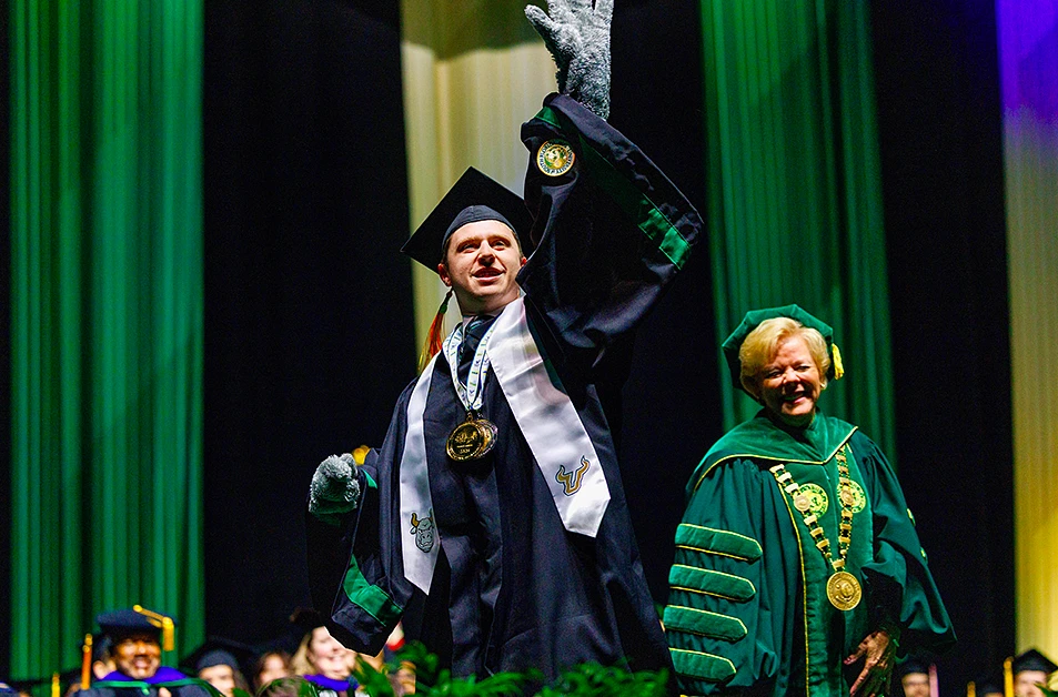 A graduate in cap and gown enthusiastically gestures on stage alongside a smiling academic official.