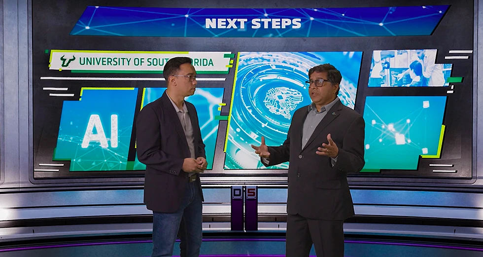 Two people are standing in front of a digital backdrop with "NEXT STEPS" and "University of South Florida" featuring AI-related graphics.